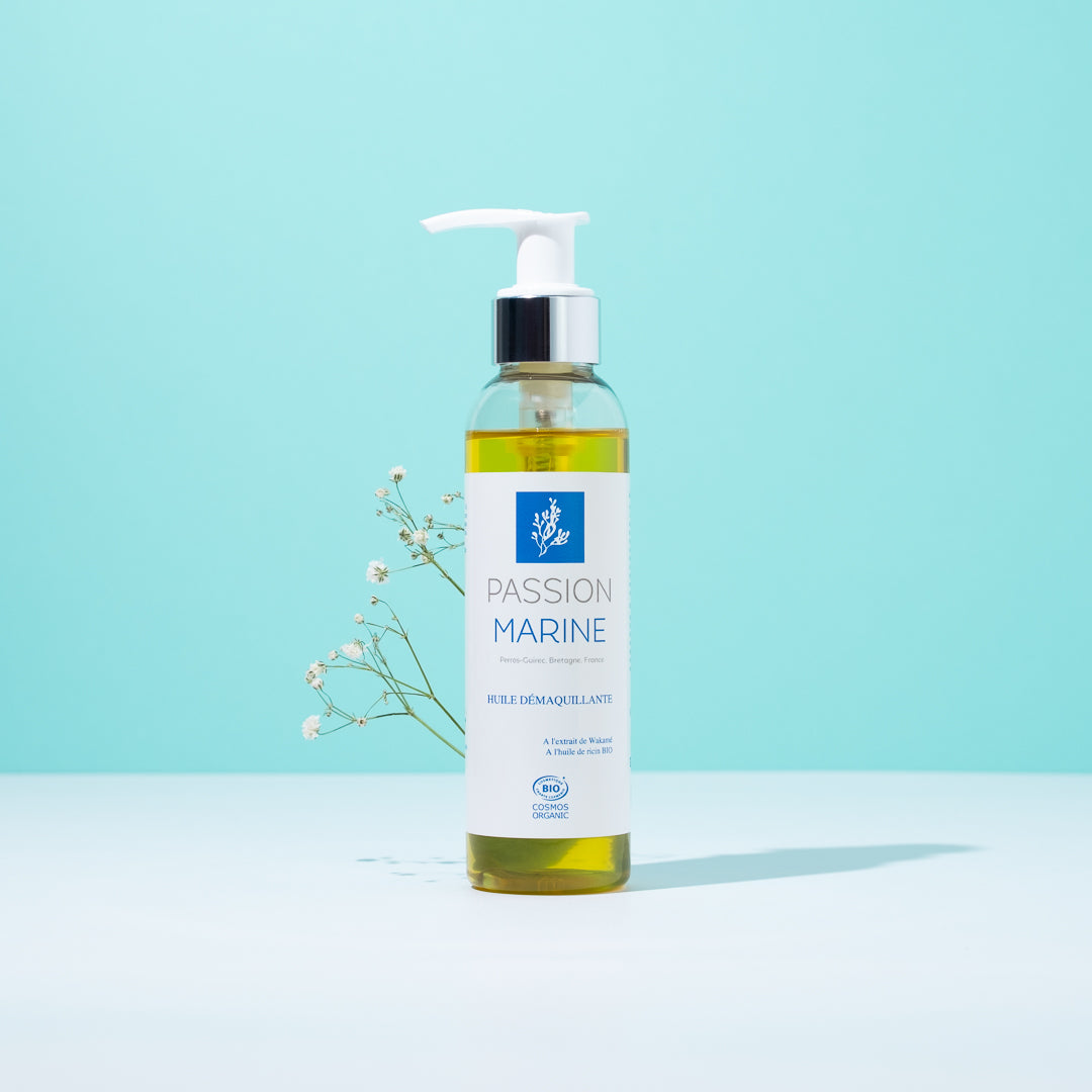 Bouteille d'huile démaquillante Passion Marine devant fond bleu avec fleurs blanches.